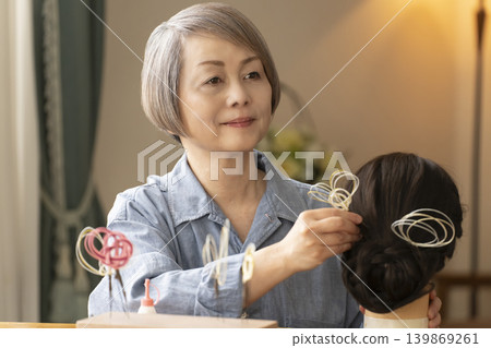 A senior woman makes a hair ornament using mizuhiki (decorative cord) and inserts it into a mannequin. A senior woman makes a hair ornament using mizuhiki (decorative cord) and inserts it into a mannequin. 139869261