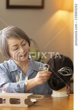 一位老婦人用水引繩製作髮飾,並將其插入人體模型中。 一位老婦人用水引繩製作髮飾,並將其插入人體模型中。 139869262