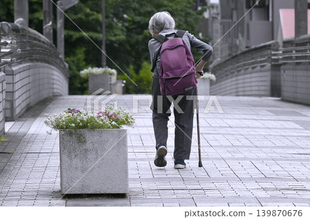 A cityscape of Yokohama, Japan: An elderly woman with a bent back and a cane, carrying a backpack... She has difficulty using the stairs of a pedestrian overpass and has to take a detour. A cityscape of Yokohama, Japan: An elderly woman with a bent back and a cane, carrying a backpack... She has difficulty using the stairs of a pedestrian overpass and has to take a detour. 139870676