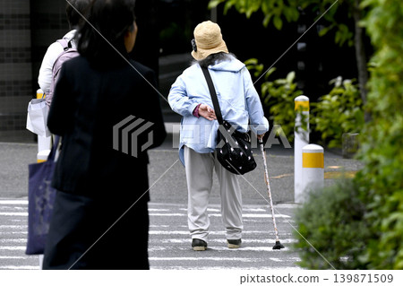 A street scene in Yokohama, Japan: An elderly woman with a cane and a bent back crosses the street... accompanied by a kind-looking woman to her left. (Yokohama City) 139871509
