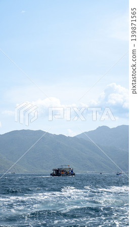 Traditional Vietnamese tour boat sailing on choppy blue sea with green mountain islands in the background. Clear sky, white waves, and distant boats near Nha Trang, Vietnam. Traditional Vietnamese tour boat sailing on choppy blue sea with green mountain islands in the background. Clear sky, white waves, and distant boats near Nha Trang, Vietnam. 139871565