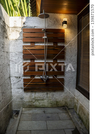 Outdoor shower area in a luxury tropical resort in Vietnam. Rain shower head on teak wood panel, stone walls, wooden ceiling, wall lamps, and lush tropical plants overhead. Outdoor shower area in a luxury tropical resort in Vietnam. Rain shower head on teak wood panel, stone walls, wooden ceiling, wall lamps, and lush tropical plants overhead. 139871570