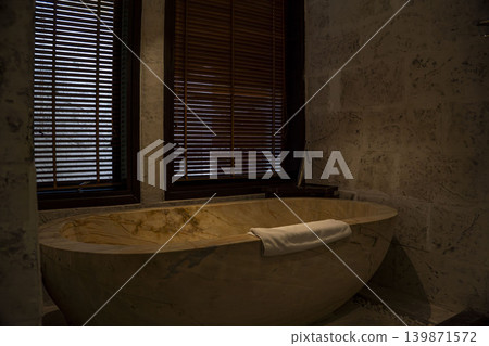 Stunning freestanding onyx stone bathtub with golden veining and white towel in a luxury resort bathroom in Vietnam. Dark wood blinds, marble walls, and warm ambient lighting. Stunning freestanding onyx stone bathtub with golden veining and white towel in a luxury resort bathroom in Vietnam. Dark wood blinds, marble walls, and warm ambient lighting. 139871572
