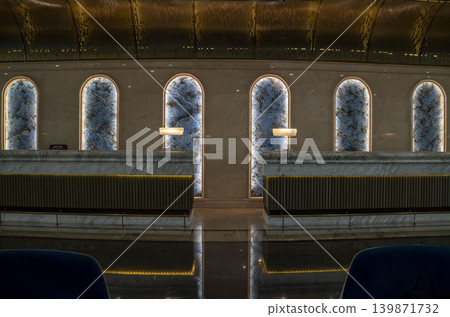 Symmetrical luxury reception area with backlit blue onyx arched panels, double marble counters, gold perforated ceiling, and elegant desk lamps. Dramatic and glamorous grand interior. Symmetrical luxury reception area with backlit blue onyx arched panels, double marble counters, gold perforated ceiling, and elegant desk lamps. Dramatic and glamorous grand interior. 139871732