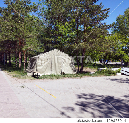 Large canvas camping tent set up in a green park surrounded by tall pine trees on a sunny day. Outdoor temporary shelter near a paved walkway in a peaceful natural environment 139872944
