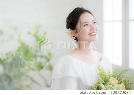 A smiling Japanese woman holding a bouquet of flowers by a window bathed in natural light, evoking images of a bride or bridal; looking up, gazing. A smiling Japanese woman holding a bouquet of flowers by a window bathed in natural light, evoking images of a bride or bridal; looking up, gazing. 139873084
