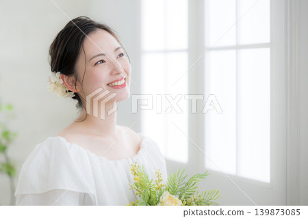 一位微笑的日本女子手捧一束鮮花,站在沐浴著自然光的窗邊,讓人聯想到新娘或待嫁女子;她抬頭凝視著窗外。 一位微笑的日本女子手捧一束鮮花,站在沐浴著自然光的窗邊,讓人聯想到新娘或待嫁女子;她抬頭凝視著窗外。 139873085