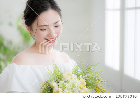 一位日本女子手捧鮮花,站在沐浴著自然光的窗邊,面帶微笑,宛如新娘或待嫁女子;她低著頭。 一位日本女子手捧鮮花,站在沐浴著自然光的窗邊,面帶微笑,宛如新娘或待嫁女子;她低著頭。 139873086