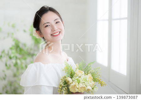 A Japanese woman smiles and looks at the camera, holding a bouquet of flowers, by a window bathed in natural light; a bride or bridal image. A Japanese woman smiles and looks at the camera, holding a bouquet of flowers, by a window bathed in natural light; a bride or bridal image. 139873087