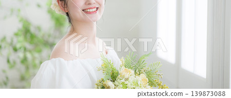 一位日本女子麵帶微笑,手捧鮮花,站在沐浴著自然光的窗前,看向鏡頭;橫幅上沒有她的臉,暗示著新娘或婚紗的形象。 一位日本女子麵帶微笑,手捧鮮花,站在沐浴著自然光的窗前,看向鏡頭;橫幅上沒有她的臉,暗示著新娘或婚紗的形象。 139873088