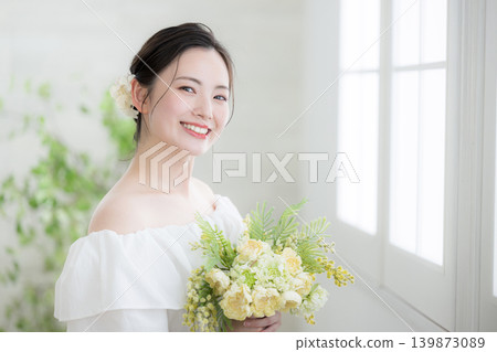 A Japanese woman smiling and looking at the camera, holding a bouquet of flowers, by a window in natural light; a bride or bridal image. Copy space on the left. A Japanese woman smiling and looking at the camera, holding a bouquet of flowers, by a window in natural light; a bride or bridal image. Copy space on the left. 139873089