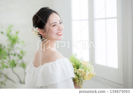 一位微笑的日本女子手捧一束鮮花,站在沐浴著自然光的窗邊,讓人聯想到新娘或待嫁女子;她轉過身,側臉示人。 一位微笑的日本女子手捧一束鮮花,站在沐浴著自然光的窗邊,讓人聯想到新娘或待嫁女子;她轉過身,側臉示人。 139873093
