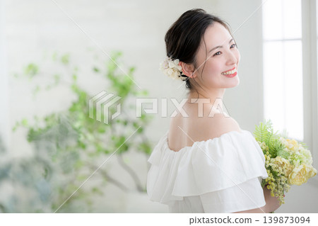 一位微笑的日本女子手捧一束鮮花,站在沐浴著自然光的窗邊,讓人聯想到新娘或待嫁女子;她轉過身,側臉示人。 一位微笑的日本女子手捧一束鮮花,站在沐浴著自然光的窗邊,讓人聯想到新娘或待嫁女子;她轉過身,側臉示人。 139873094