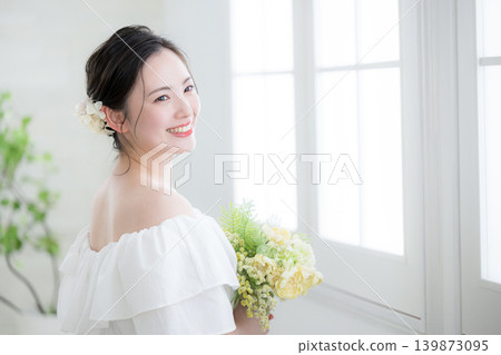 A smiling Japanese woman holding a bouquet of flowers by a window bathed in natural light, evoking images of a bride or bridal; looking up, gazing. A smiling Japanese woman holding a bouquet of flowers by a window bathed in natural light, evoking images of a bride or bridal; looking up, gazing. 139873095