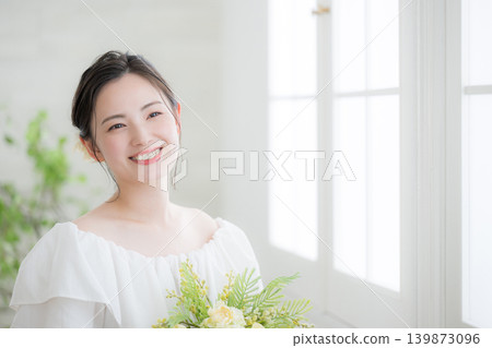 一位日本女子麵帶微笑,手捧一束鮮花,站在沐浴著自然光的窗邊,看向鏡頭;這是一張新娘或婚紗照。 一位日本女子麵帶微笑,手捧一束鮮花,站在沐浴著自然光的窗邊,看向鏡頭;這是一張新娘或婚紗照。 139873096