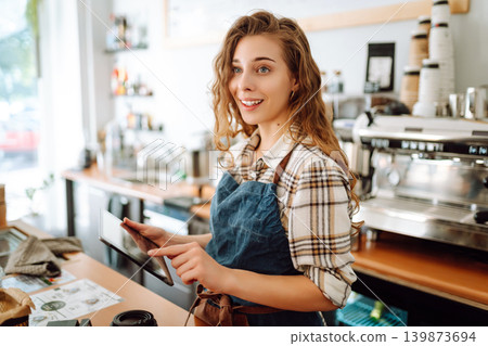 A young coffee shop owner standing behind bar using a digital tablet. Takeaway concept, technology. A young coffee shop owner standing behind bar using a digital tablet. Takeaway concept, technology. 139873694