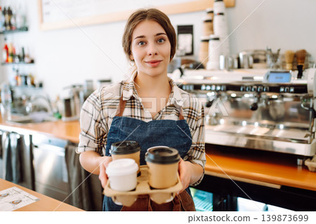The young barista owner behind the bar gives out to-go orders. Business concept. Drinks to go. The young barista owner behind the bar gives out to-go orders. Business concept. Drinks to go. 139873699
