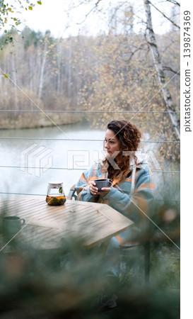 A sweet woman with curly hair drinks tea and relaxes on the terrace of her house on an autumn day. Health care, sincerity, a sense of balance and calm. 139874369