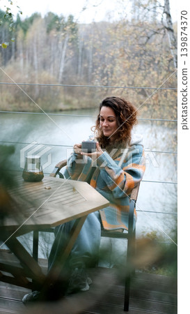 A sweet woman with curly hair drinks tea and relaxes on the terrace of her house on an autumn day. Health care, sincerity, a sense of balance and calm. 139874370