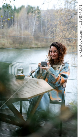 A sweet woman with curly hair drinks tea and relaxes on the terrace of her house on an autumn day. Health care, sincerity, a sense of balance and calm. 139874371