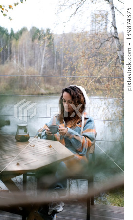 A sweet woman with curly hair drinks tea and relaxes on the terrace of her house on an autumn day. Health care, sincerity, a sense of balance and calm. A sweet woman with curly hair drinks tea and relaxes on the terrace of her house on an autumn day. Health care, sincerity, a sense of balance and calm. 139874375
