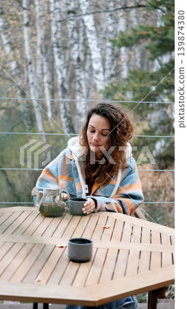 A sweet woman with curly hair drinks tea and relaxes on the terrace of her house on an autumn day. Health care, sincerity, a sense of balance and calm. A sweet woman with curly hair drinks tea and relaxes on the terrace of her house on an autumn day. Health care, sincerity, a sense of balance and calm. 139874380
