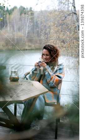 A sweet woman with curly hair drinks tea and relaxes on the terrace of her house on an autumn day. Health care, sincerity, a sense of balance and calm. 139874398