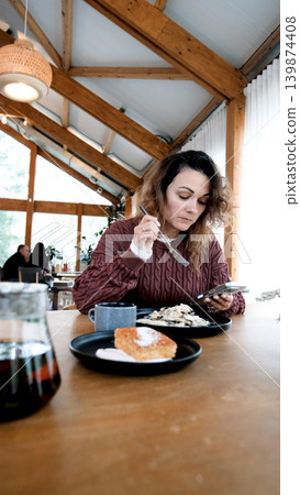A young woman eats pasta in a restaurant and texts on her smartphone. 139874408
