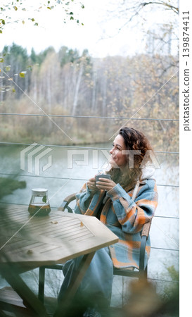 A sweet woman with curly hair drinks tea and relaxes on the terrace of her house on an autumn day. Health care, sincerity, a sense of balance and calm. A sweet woman with curly hair drinks tea and relaxes on the terrace of her house on an autumn day. Health care, sincerity, a sense of balance and calm. 139874411