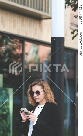 Caucasian woman with curly hair using her mobile phone outdoors. Close-up. Communication technology and modern lifestyle concept Caucasian woman with curly hair using her mobile phone outdoors. Close-up. Communication technology and modern lifestyle concept 139874422