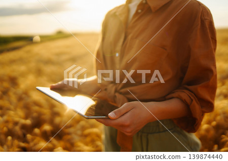 Young woman stands in field of wheat as sun sets. She holds a document, smiles. Young woman stands in field of wheat as sun sets. She holds a document, smiles. 139874440