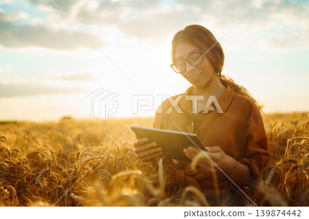 Young woman stands in field of wheat as sun sets. She holds a document, smiles. Young woman stands in field of wheat as sun sets. She holds a document, smiles. 139874442