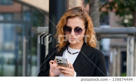 Caucasian woman with curly hair using her mobile phone outdoors. Close-up. Communication technology and modern lifestyle concept Caucasian woman with curly hair using her mobile phone outdoors. Close-up. Communication technology and modern lifestyle concept 139874494