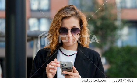 Caucasian woman with curly hair using her mobile phone outdoors. Close-up. Communication technology and modern lifestyle concept Caucasian woman with curly hair using her mobile phone outdoors. Close-up. Communication technology and modern lifestyle concept 139874495