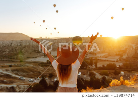 Woman stands on mountain with flying air balloons on background. Tourist Turkish region Cappadocia. Woman stands on mountain with flying air balloons on background. Tourist Turkish region Cappadocia. 139874714
