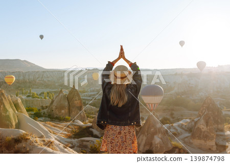 Happy girl in hat stands on mountain with flying air balloons on background. View from the back. Happy girl in hat stands on mountain with flying air balloons on background. View from the back. 139874798