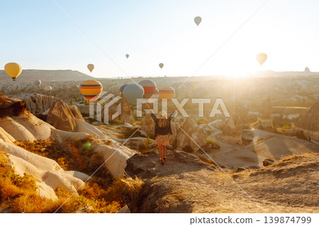 Happy girl in hat stands on mountain with flying air balloons on background. View from the back. Happy girl in hat stands on mountain with flying air balloons on background. View from the back. 139874799