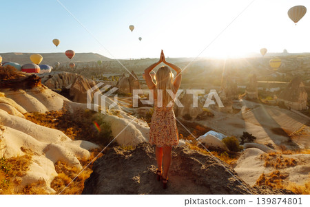 Happy girl in hat stands on mountain with flying air balloons on background. View from the back. 139874801