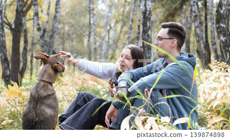 A married couple relaxes in nature during a walk with their beloved dog in an autumn forest. The loving woman and man sit in the tall yellow grass, in the fern leaves, with the dog next to them. A married couple relaxes in nature during a walk with their beloved dog in an autumn forest. The loving woman and man sit in the tall yellow grass, in the fern leaves, with the dog next to them. 139874898