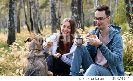 A married couple relaxes in nature during a walk with their beloved dog in an autumn forest. The loving woman and man sit in the tall yellow grass, in the fern leaves, with the dog next to them. A married couple relaxes in nature during a walk with their beloved dog in an autumn forest. The loving woman and man sit in the tall yellow grass, in the fern leaves, with the dog next to them. 139874965