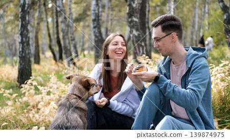 A married couple relaxes in nature during a walk with their beloved dog in an autumn forest. The loving woman and man sit in the tall yellow grass, in the fern leaves, with the dog next to them. 139874975