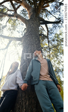 Lovers boy and girl standing near a big pine tree during a walk on a sunny autumn day Lovers boy and girl standing near a big pine tree during a walk on a sunny autumn day 139874983