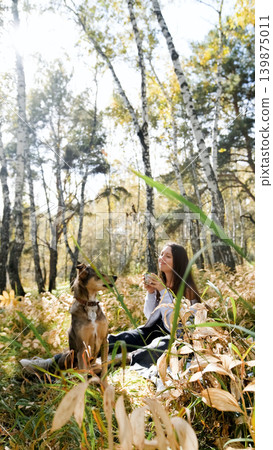 A sweet young woman with her dog enjoys the beauty of nature and the bond with her pets. The woman drinks tea from a thermos and eats buns A sweet young woman with her dog enjoys the beauty of nature and the bond with her pets. The woman drinks tea from a thermos and eats buns 139875011