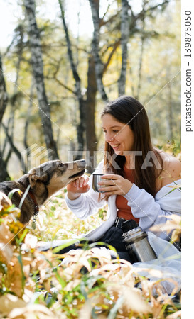 A sweet young woman with her dog enjoys the beauty of nature and the bond with her pets. The woman drinks tea from a thermos A sweet young woman with her dog enjoys the beauty of nature and the bond with her pets. The woman drinks tea from a thermos 139875050