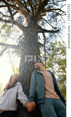 Lovers boy and girl standing near a big pine tree during a walk on a sunny autumn day Lovers boy and girl standing near a big pine tree during a walk on a sunny autumn day 139875582