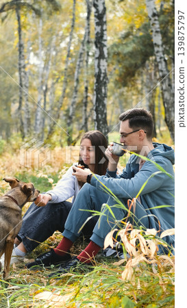 A married couple relaxes in nature during a walk with their beloved dog in an autumn forest. The loving woman and man sit in the tall yellow grass, in the fern leaves, with the dog next to them. 139875797