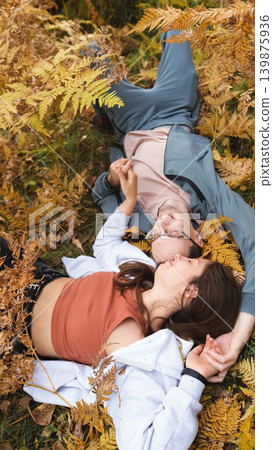 A woman and a man in love lie in tall yellow grass, in fern leaves. The married couple relaxes in nature during a walk in the autumn forest. A woman and a man in love lie in tall yellow grass, in fern leaves. The married couple relaxes in nature during a walk in the autumn forest. 139875936