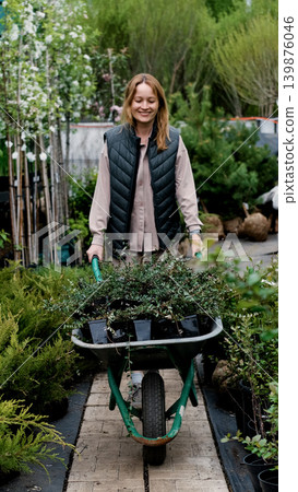 A female gardener carries plant seedlings in a cart at a nursery or garden center. A female gardener carries plant seedlings in a cart at a nursery or garden center. 139876046