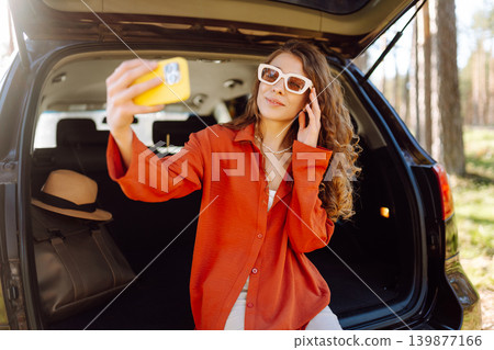 A cheerful woman sits on the trunk of her car and taking a selfie. Selfie while traveling by car. 139877166