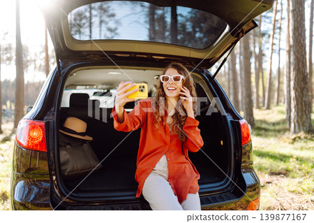 A cheerful woman sits on the trunk of her car and taking a selfie. Selfie while traveling by car. A cheerful woman sits on the trunk of her car and taking a selfie. Selfie while traveling by car. 139877167